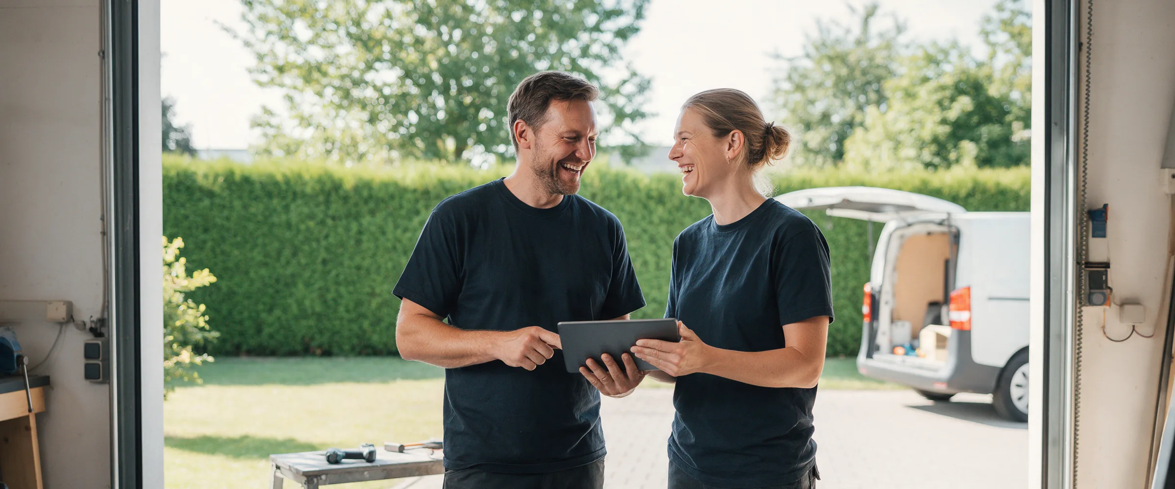 Zwei Handwerker stehen im offenen Tor einer Werkstatt und sehen gemeinsam auf ein Tablet, dahinter der Hof mit Hecke und Transporter