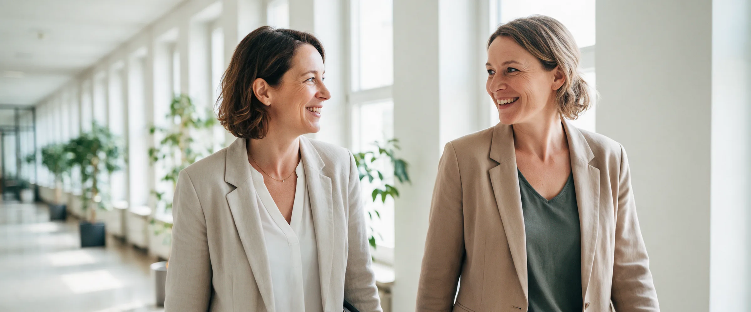 Zwei Frauen gehen im Gespräch durch den hellen Flur eines Verwaltungsgebäudes, eine von ihnen trägt eine Aktenmappe