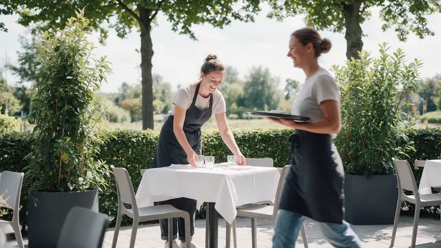 Auf der begrünten Terrasse eines Cafés deckt eine Mitarbeiterin einen Tisch ein, eine zweite geht mit einem Tablett vorbei